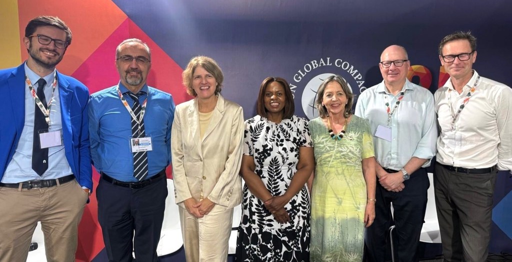 Está foto muestra a un grupo diverso de seis delegados en lo que parece ser un panel de discusión o un diálogo estratégico.
• Delegados: Cuatro hombres y dos mujeres están de pie en una fila.
• La mujer en el centro, vestida de blanco y beige claro, es una de las figuras clave de la delegación de Pacto Global Chile,  Margarita Ducci, Directora Ejecutiva, dada su presencia y mención destacada en la cobertura de Pacto Global sobre la COP30.
• Fondo y Contexto: El fondo es de color oscuro y vibrante, con el logo o texto de "UN GLOBAL COMPAC..." (Pacto Global de las Naciones Unidas), visible en el centro. Esto sugiere que el panel está directamente relacionado con las actividades de la Red Pacto Global de las Naciones Unidas a nivel internacional, enfocándose en la agenda climática global y la aceleración de la acción empresarial.
• Ambiente: El vestuario, predominantemente formal (trajes y corbatas en los hombres), sugiere que se trata de un Diálogo de Alto Nivel con la participación de figuras internacionales y líderes de la ONU, lo que reafirma el rol de Pacto Global Chile en la articulación de esfuerzos con el Sistema de Naciones Unidas y el gobierno chileno de cara a la COP30.